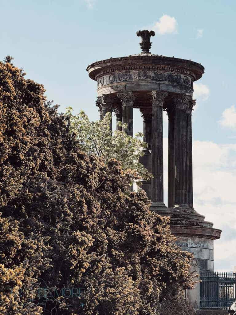 A tall stone monument with columns stands high above stone walls and shrubs against a cloudy blue sky. This is Calton Hill in Edinburgh.