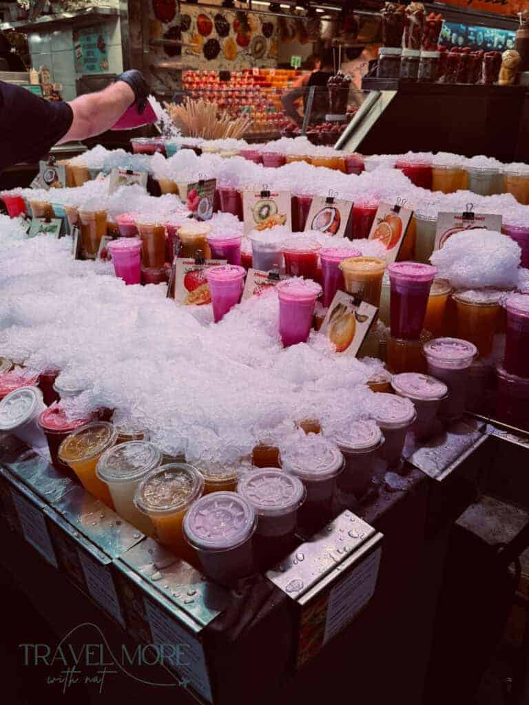 Market stall filled with colourful fruit juices and smoothies displayed in cups surrounded by crushed ice. Refreshing cold drinks in shades of pink, orange and purple highlight the importance of hydration when using heatwave travel tips during summer trips.