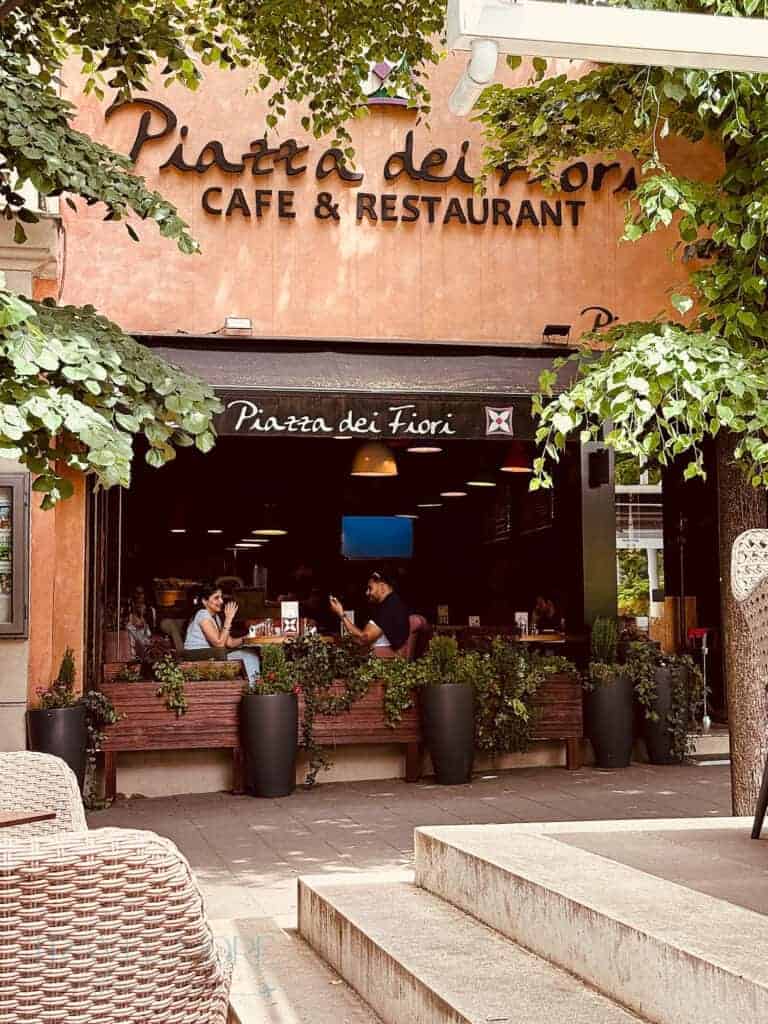 Outdoor seating area at Piazza dei Fiori Cafe and Restaurant shaded by leafy trees on a sunny day. Diners relax in the cooler shade, showing a practical way to enjoy meals outside while staying comfortable during extreme summer temperatures.