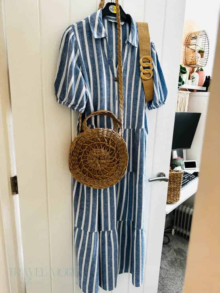 Blue and white striped midi dress hanging on a door with a woven round handbag and straw belt beside it. Lightweight clothing and breathable accessories shown as outfit inspiration for staying comfortable during hot weather and following heatwave travel tips.