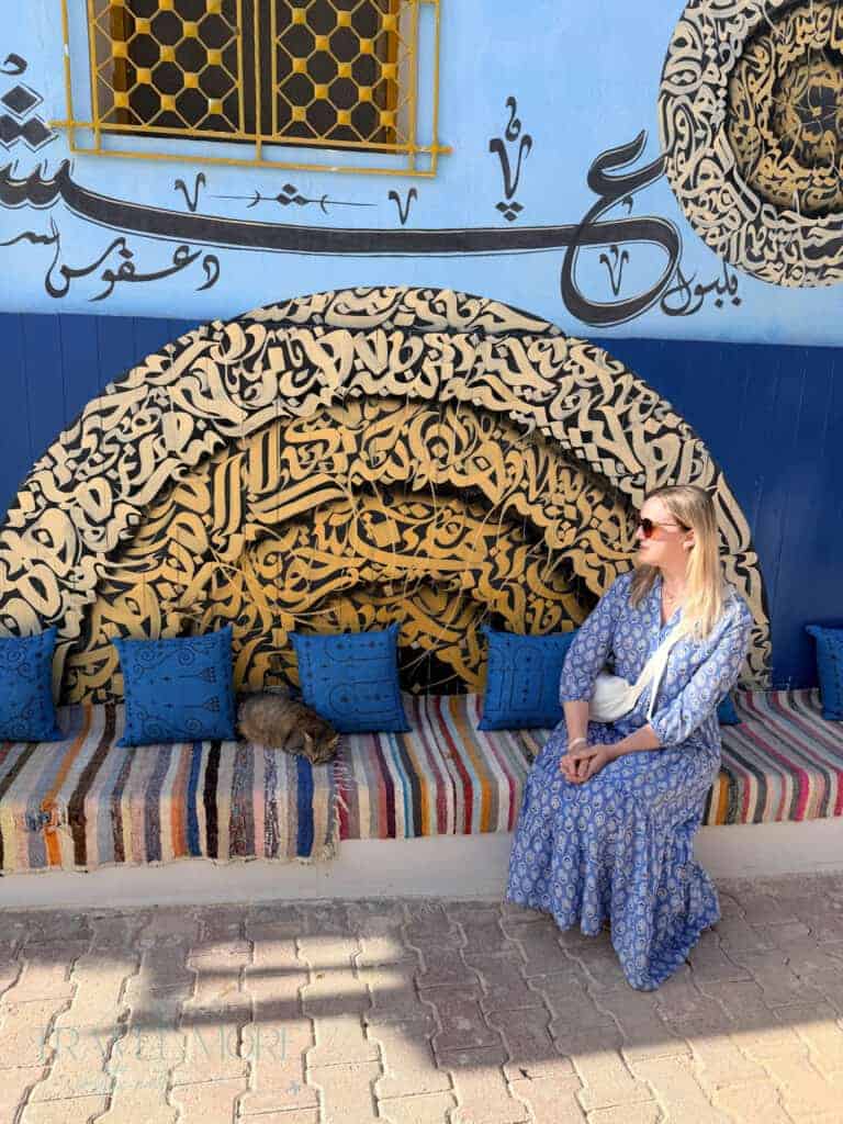 Woman in a blue patterned dress sitting beside a striped bench beneath an Arabic calligraphy mural.