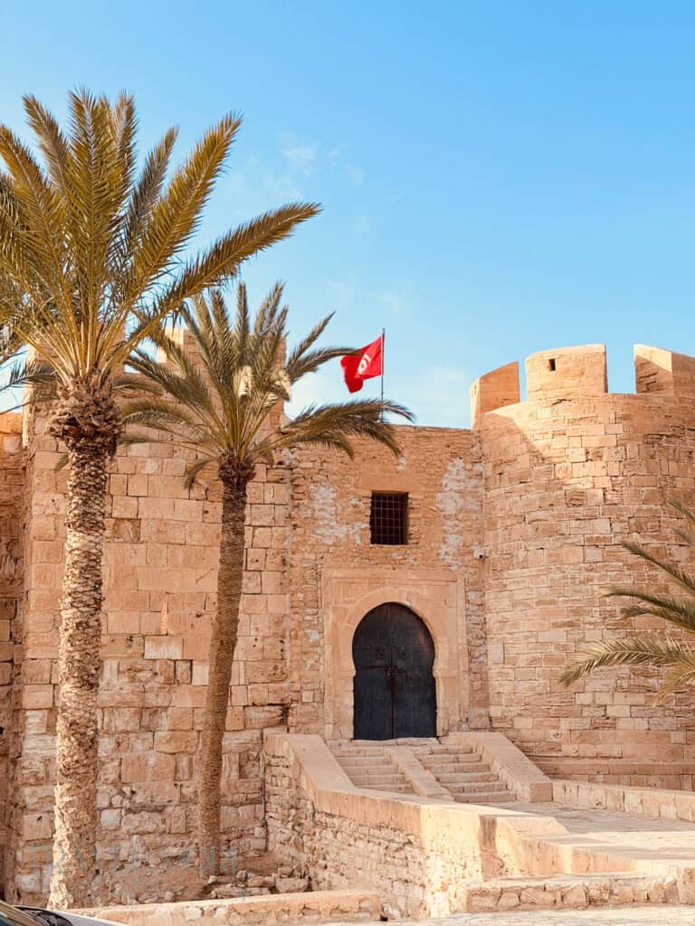 Palm trees and a red Tunisian flag frame a historic stone fort in Djerba, Tunisia - a must-see during any Djerba itinerary.