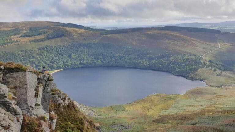 A lone hiker sits on a rocky ledge overlooking Lough Tay, surrounded by dense forests and rolling hills. The lake's dark waters contrast with the vibrant greenery under a cloudy sky.