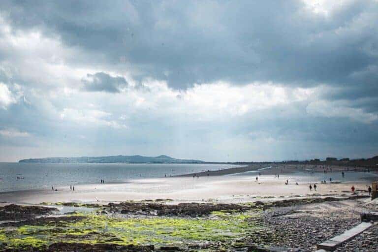 A wide view of the beach stretching from Portmanock to Malahide, Ireland. The foreground includes green algae on rocks, with people walking along the expansive shoreline under a cloudy sky.