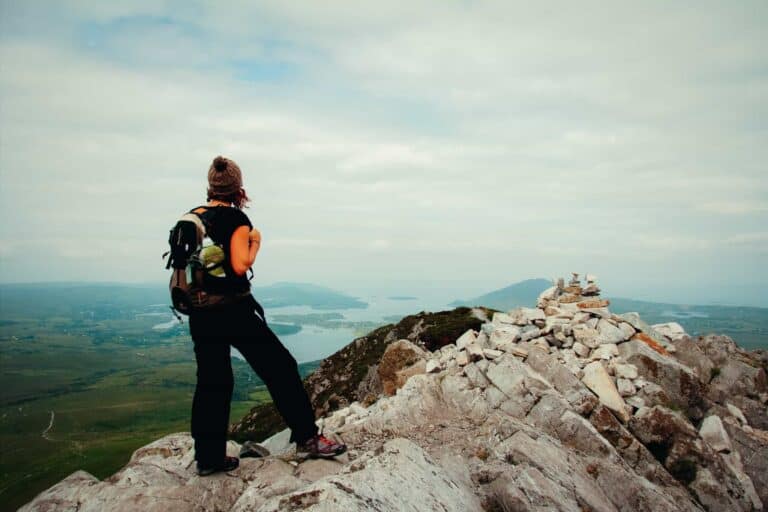 A hiker standing on the rocky summit of Diamond Hill in Connemara, overlooking a scenic view of lakes and hills.