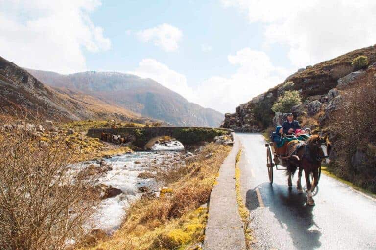 A horse-drawn carriage carrying people along a winding mountain road in the Gap of Dunloe, with a river and stone bridge in the background under a bright sky.