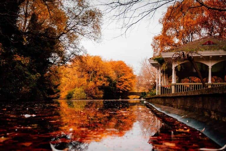 A calm autumn scene with a pond reflecting fallen red and orange leaves, and a stone bridge in the background. The gazebo on the right overlooks the water, with trees in vibrant fall colors adding to the serene atmosphere.