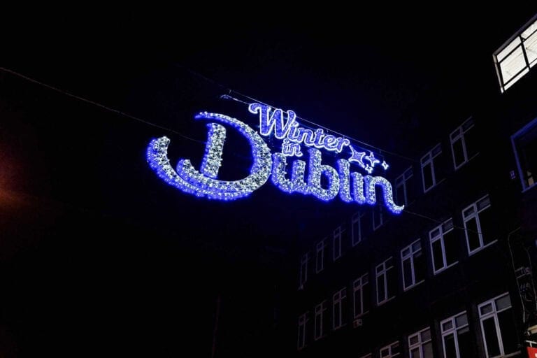 A glowing street sign reading "Winter in Dublin" adorned with bright white and blue lights, hanging against a dark night sky, with a building faintly visible in the background.