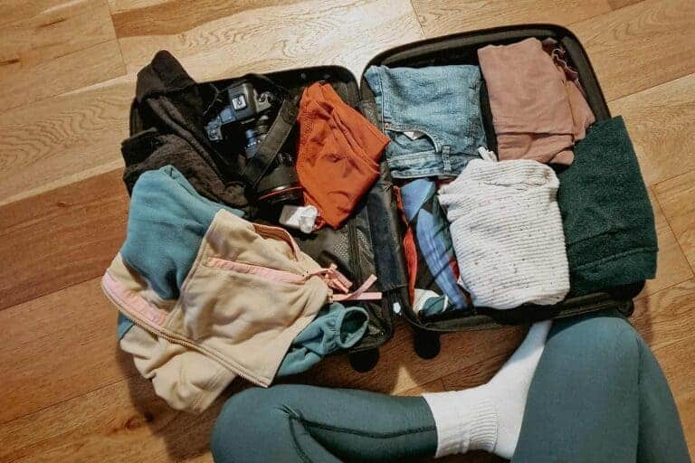An open suitcase packed with clothing and essentials, including a fleece jacket, jeans, folded sweaters, a plaid scarf, and a camera with a lens. The suitcase sits on a wooden floor, with part of a person's leg and foot in socks visible in the frame, suggesting preparation for a trip.