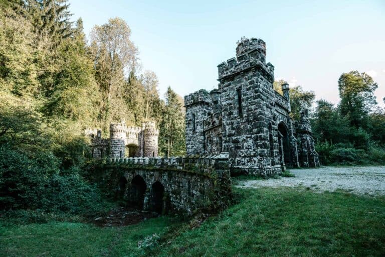 A forest pathway leading through the arched gateway of Ballysaggartmore Towers, framed by rugged stonework and vibrant green trees in the background.