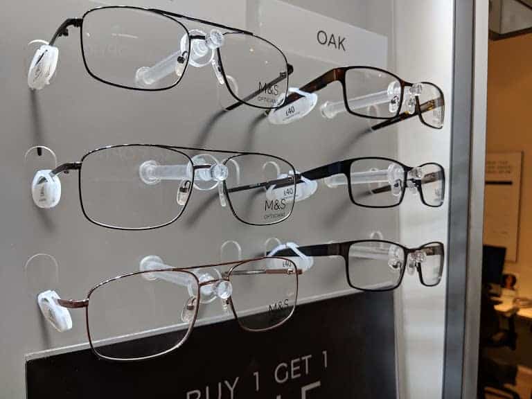 Display of six pairs of eyeglasses on a wall, labeled "OAK," reminiscent of the quality seen at Marks and Spencer Opticians. Signs with pricing and a "Buy 1 Get 1 Half Price" offer are visible, inviting customers to explore exceptional eyewear deals.