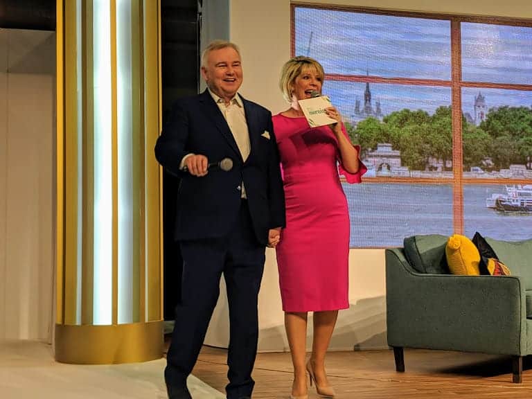 Eamonn Holmes and Ruth Langsford standing on stage at This Morning Live. Ruth is in a stunning bright pink fitted dress and Eamonn in a navy suit. Both have Microphones and smiling.