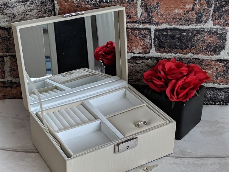 White jewellery box that is open on a sideboard, the box shows two empty trays and a ring compartment. next to the jewellery box is a plant of red flowers
