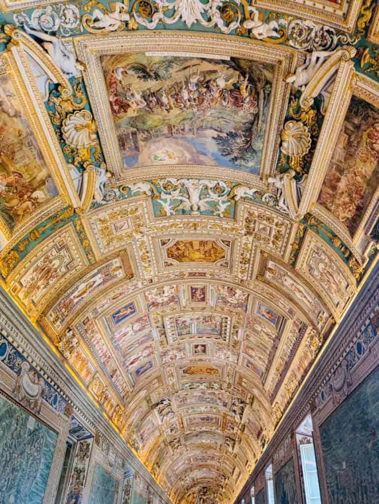 Ornate vaulted ceiling in the Gallery of Maps at the Vatican Museums, covered in detailed frescoes, gilded moldings, and religious iconography stretching down a long hallway.