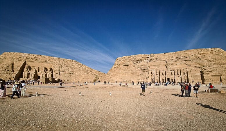 This image shows a wide-angle view of a historic Egyptian monument, the Temple of Ramses II at Abu Simbel. The temple's facade is adorned with four colossal statues of Ramses II seated on thrones, each several meters high, with smaller figures at their feet representing family members. The temple is carved directly into a solid rock cliff and the entrance is between the statues. There's a clear blue sky above and a broad, sandy area in front of the temple where visitors are scattered around, some taking photos, others walking towards the temple. The scale of the human figures compared to the statues and the temple itself emphasizes the monument's grandeur.