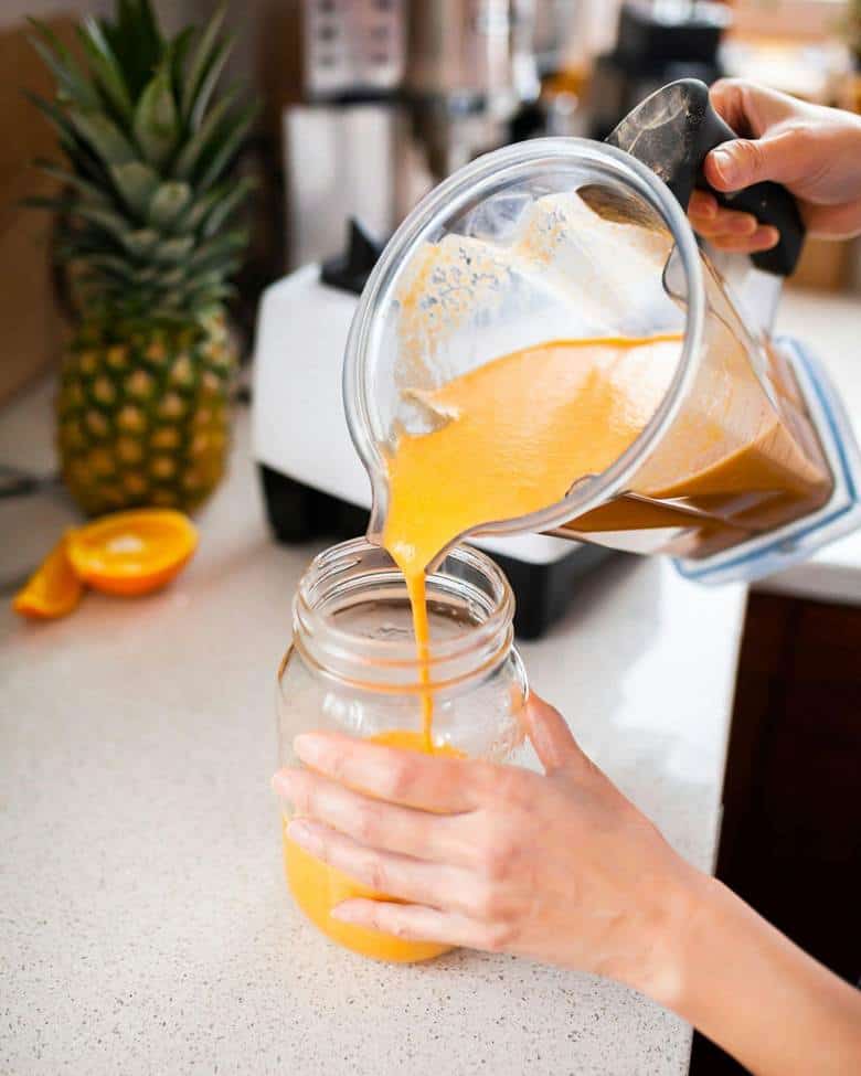 A person pours a thick orange smoothie from a blender into a mason jar on a kitchen counter, with a fresh pineapple and a halved orange nearby.