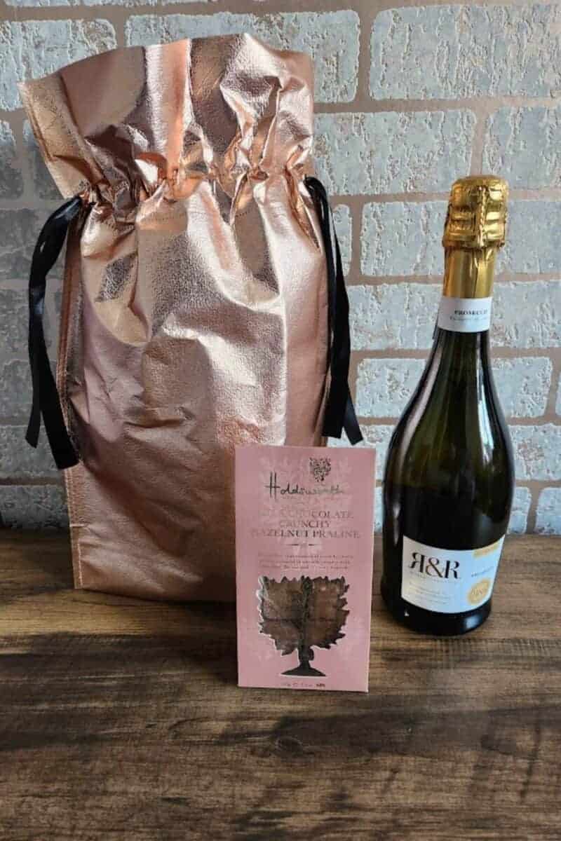 A rose gold coloured gift bag with a bottle of prosecco and a small box of hazelnut praline chocolates.