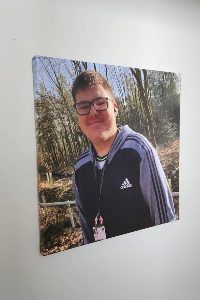 A canvas print of Ben, a teenage boy smiling in a proud and confident way whilst standing in woodland whilst wearing an adidas top, glasses and a lanyard.