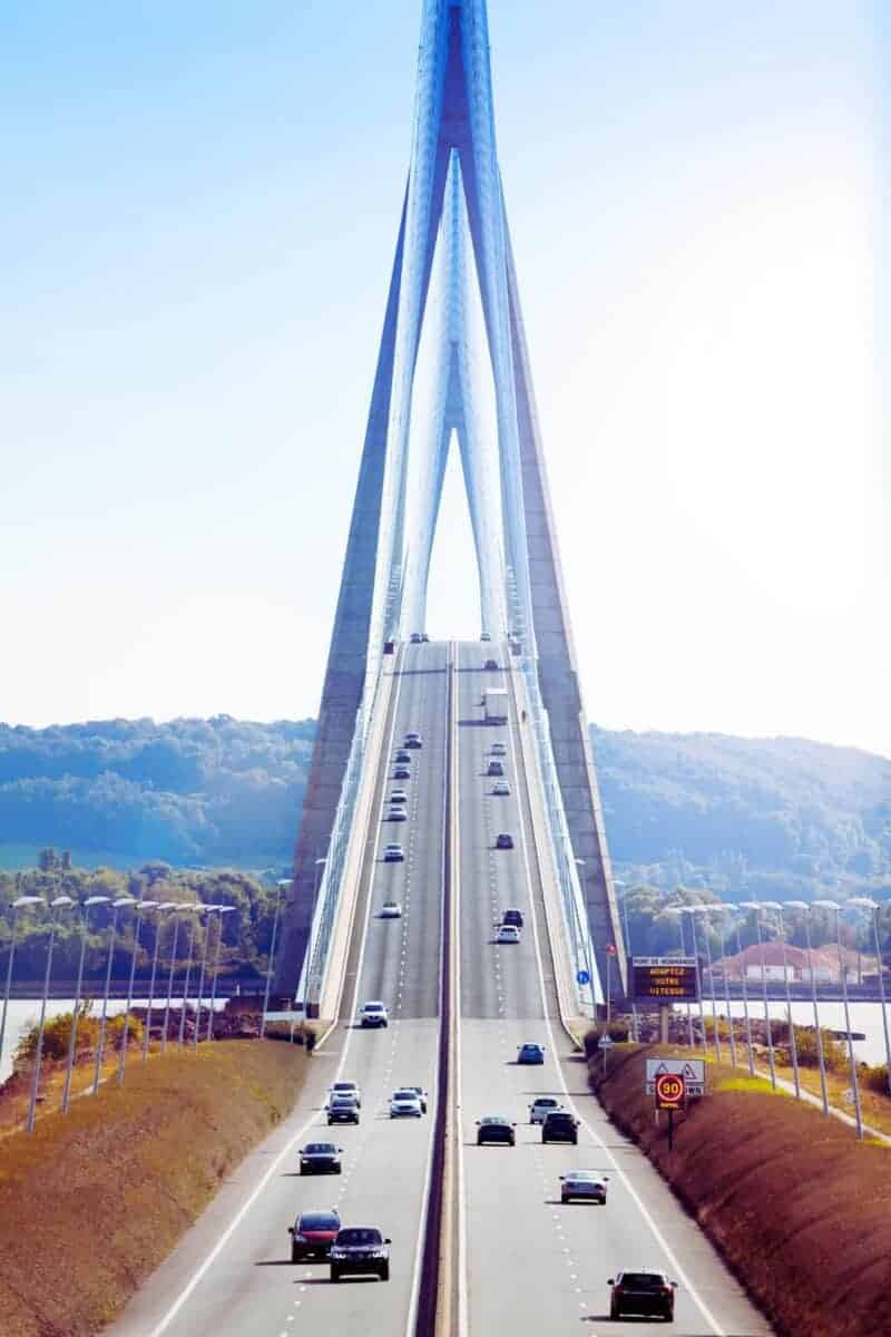 A suspension bridge with cable supports gracefully spans a river, cars traveling in both directions under a clear sky. Nearby hotels offer stunning views of this architectural marvel, providing guests an unforgettable backdrop to their stay.