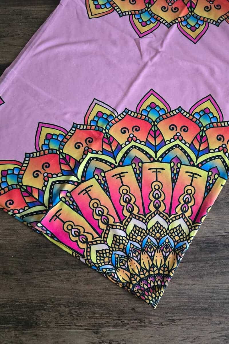 A pink fabric with vibrant mandala patterns in orange, yellow, and blue tones is spread on a wooden surface, perfect as gifts for babies with its soft and colorful appeal.