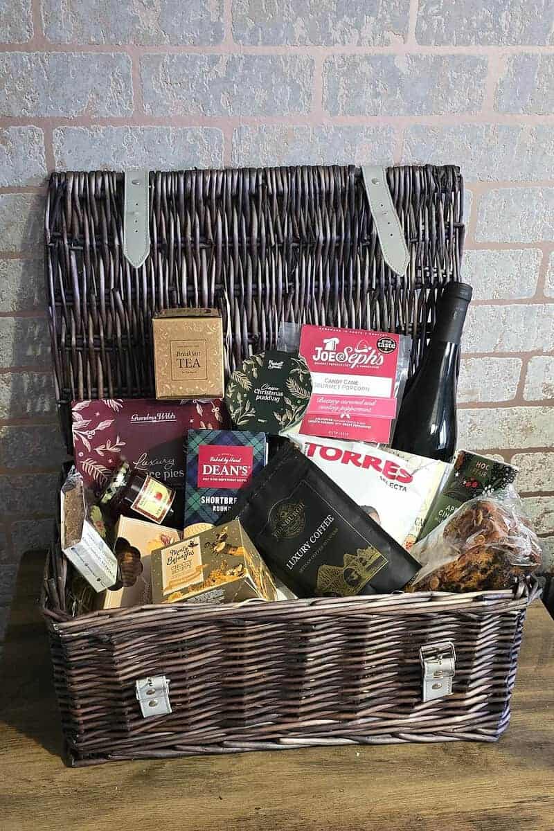 A wicker hamper, the perfect gift for families, brimming with assorted gourmet items like tea, chips, a bottle of wine, shortbread, chocolate, and snacks is set charmingly against a brick wall.
