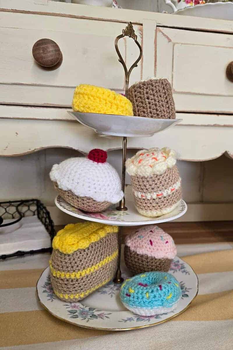 A three-tiered stand showcases crocheted desserts, like cupcakes and slices of cake, on a decorative plate against a vintage cabinet backdrop at the Sweet Spot within PoFfles Coffee and Cake Emporium.