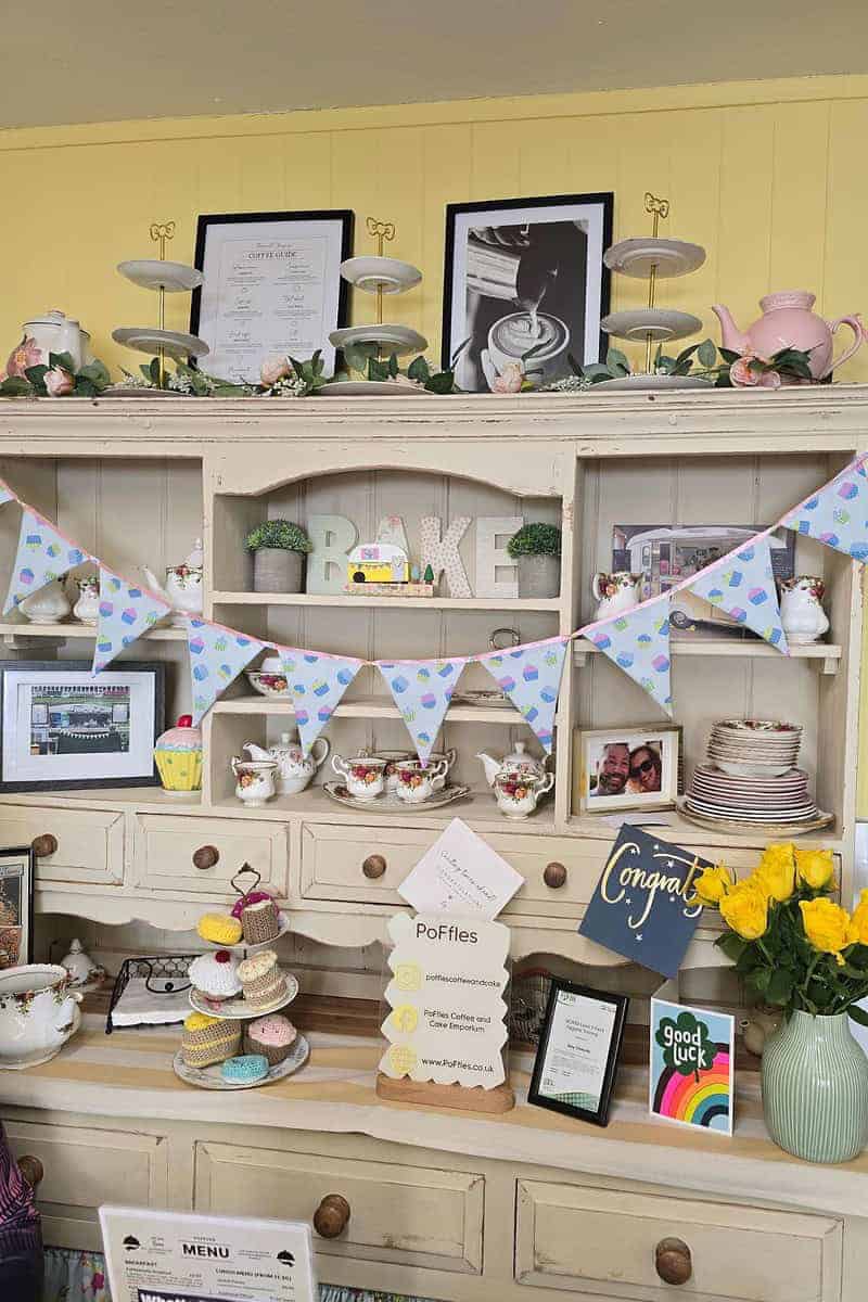 A wooden hutch at Mansfield displays colorful bunting, plates, teacups, framed photos, and decorative items. Yellow roses are placed on the right. Various signs and cards from PoFfles Coffee are arranged on the shelves.
