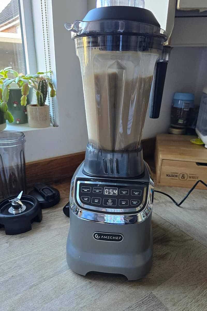 A sleek AMZCHEF Blender sits on the kitchen countertop, diligently blending a brown mixture. The digital display shows 0:54. Nearby are a plant, blender lid, and other kitchen items—a testament to the efficiency of Model 5003 in our latest Kitchen Review.