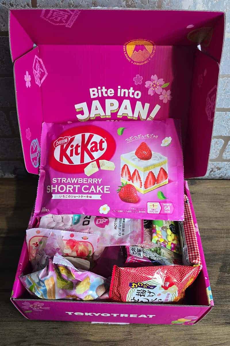 An open pink TokyoTreat box displays assorted Japanese snacks, making it a perfect Mother's Day gift. Enjoy delights like a large KitKat Strawberry Shortcake, colorful candy, and treats under a lid that reads “Bite into JAPAN!” with Mount Fuji and cherry blossoms.