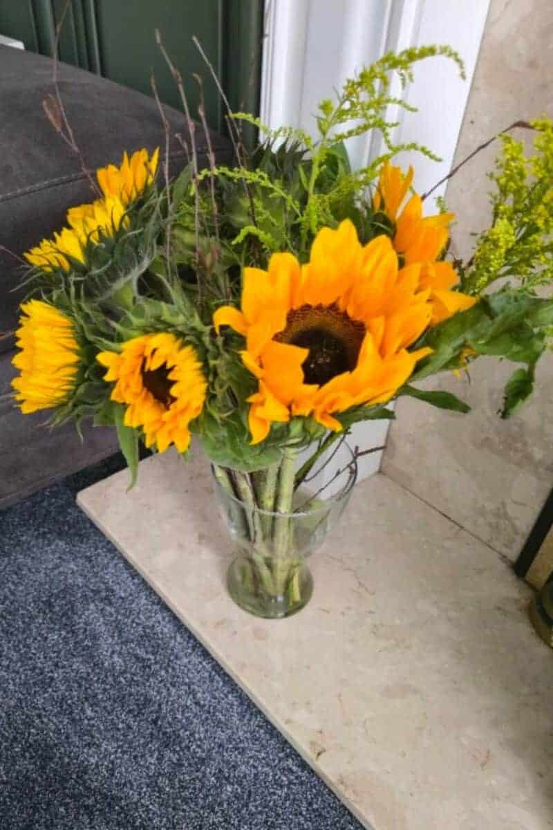 A clear glass vase holds a bouquet of bright yellow sunflowers from Prestige Flowers, with dark centers and green leaves. The arrangement sits on a marble surface by a dark couch and blue-gray carpet, against a white and green wall.