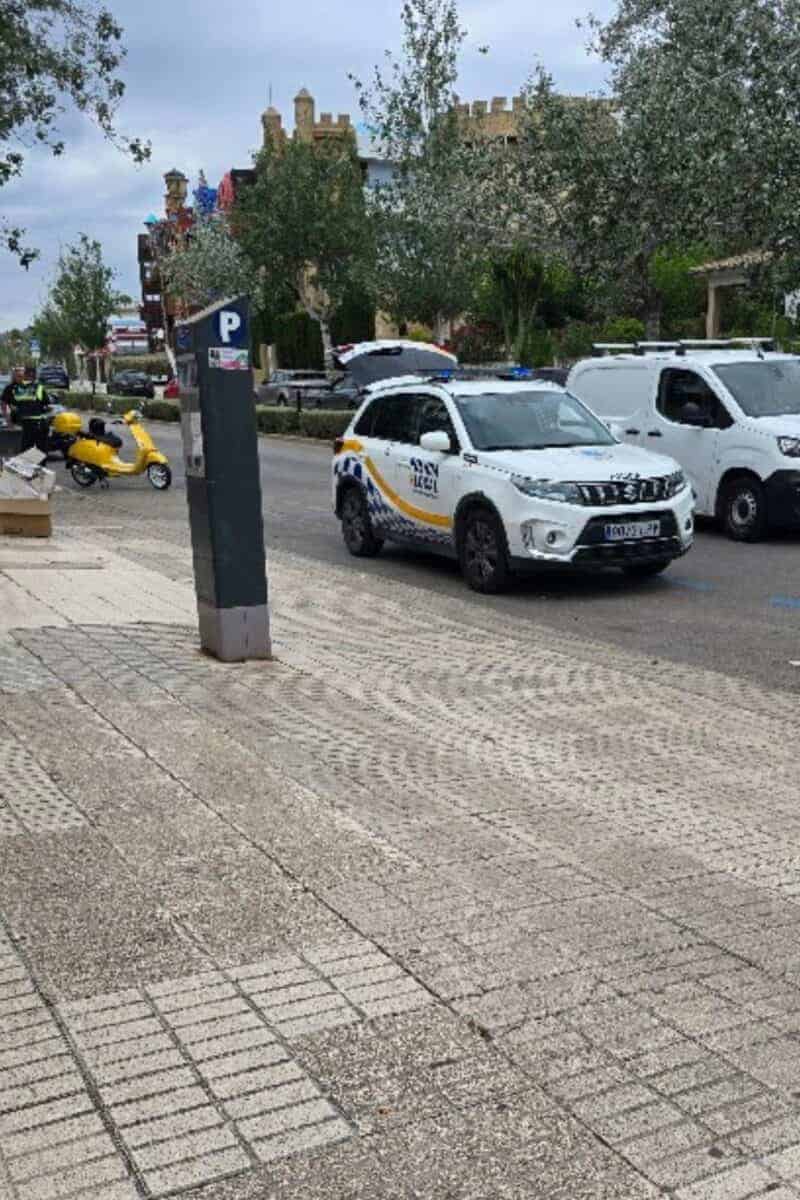 A city street scene perfect for a Mallorca driving guide: a parked traffic police SUV, yellow scooter, and white van. Pedestrians stroll beneath trees and an overcast sky, with a parking meter in the foreground—a glimpse of driving in Mallorca.