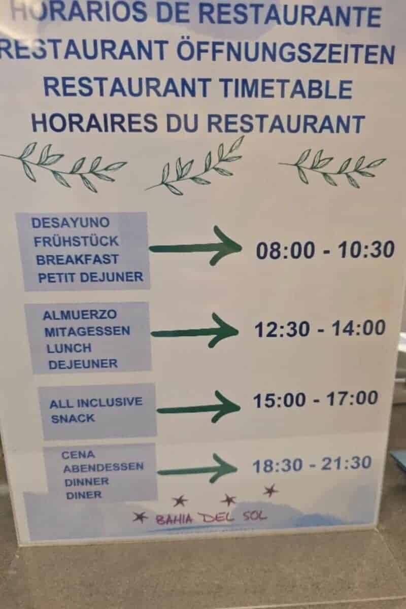 A sign at Bahia del Sol in Santa Ponsa details all-inclusive restaurant hours in four languages, listing breakfast (08:00-10:30), lunch, snack, and dinner times, with "Bahia del Sol" featured at the bottom.