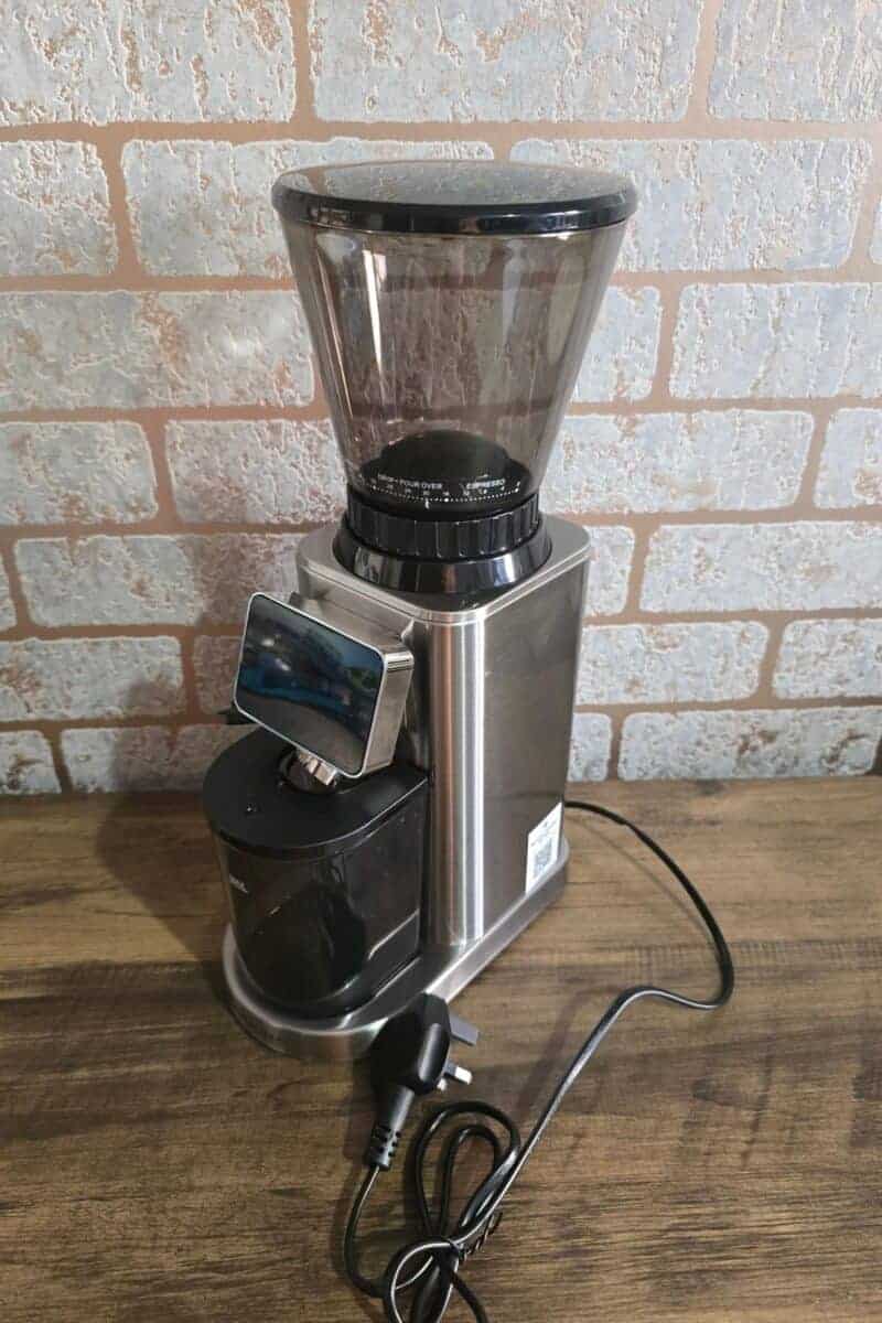 A stainless steel electric coffee grinder with a transparent bean hopper, digital display, and black grounds container sits on a wooden surface in front of a light brick wall—ready to deliver freshly ground beans every morning. The power cord is coiled in front.
