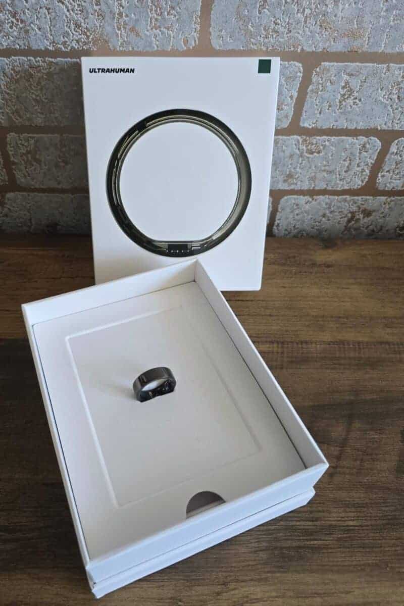 A black Ultrahuman Ring Air smart ring sits in an open white box on a wooden surface, with the product’s outer packaging labeled “Ultrahuman” standing upright in the background against a brick-patterned wall.