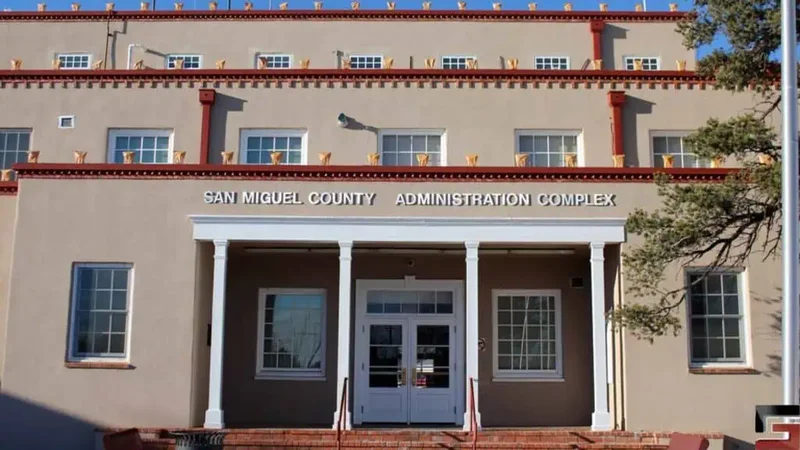 A photograph of San Miguel County Administration Complex on a sunny day. The building is tan with white and brown columns and beams.