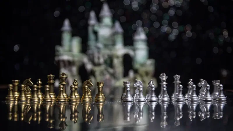 Silver and gold chess pieces are aligned on a chess board with a pewter castle blurred in the background against a black wall.