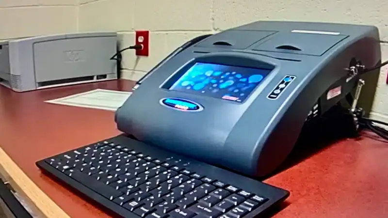 A sleek, dark grey Intoxilyzer 9000 breath alcohol testing machine sitting on a desk, featuring a color touchscreen display, an attached keyboard, and a nearby printer.
