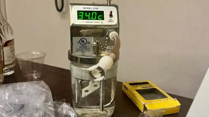 A Guth Laboratories Model 2100 wet bath simulator with a digital temperature display reading 34.02°C, connected to a glass jar filled with a clear testing solution.
