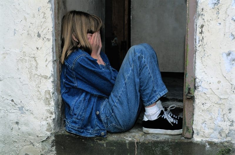 Niña rubia con chaqueta y pantalón vaqueros, sentada en el umbral de una puerta, tapándose la cara con las manos.