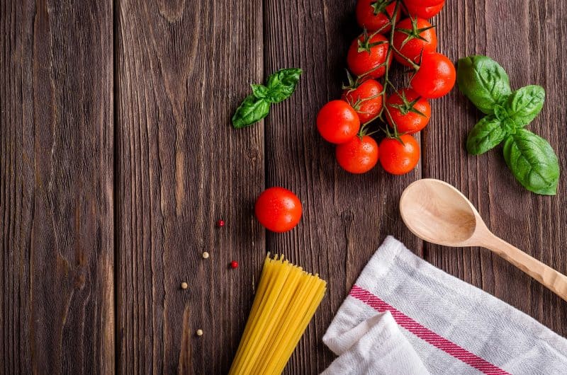 Tomates en rama, albahaca, pasta, granos de pimienta, cuchara y trapo sobre superficie de madera