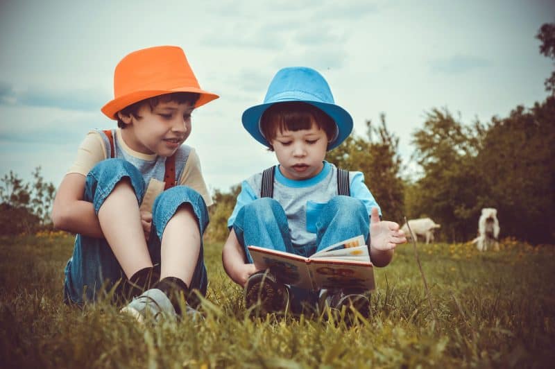 Dos niños con vaqueros y sombreros de colores vivos leen un libro sentados en el campo