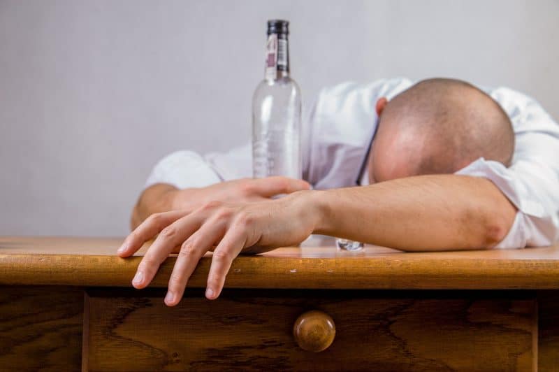 Persona recostada sobre una mesa sujetando una botella de alcohol