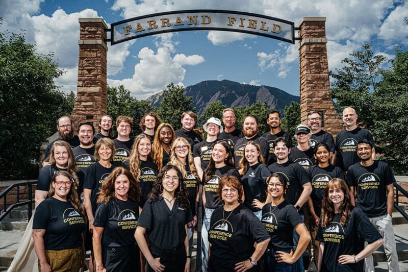 Group photo of the CU Boulder Conference Services team in black shirts, standing in front of the Farrand Field sign with mountains and blue sky in the background, illustrating their role in managing large-scale campus operations and enhancing event coordination.