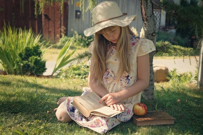 Niña rubia con vestido de estrellas y sombrero de paja, sentada junto a un árbol en el jardín, leyendo un libro. Junto a ella hay otro libro con una manzana encima.