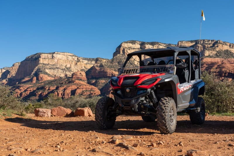 Red and gray off-road UTV on a rocky desert trail with red canyon walls in the background, clear blue sky above