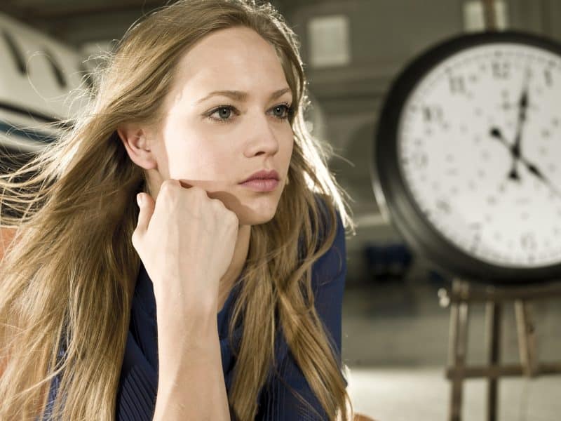 Chica rubia junto a un reloj, con la cabeza apoyada sobre su mano.