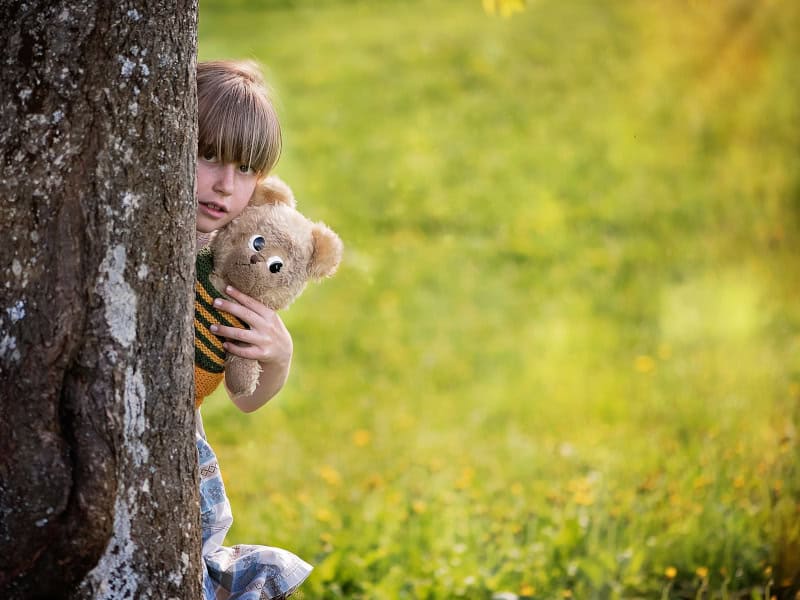 Niña con osito de peluche detrás de un árbol