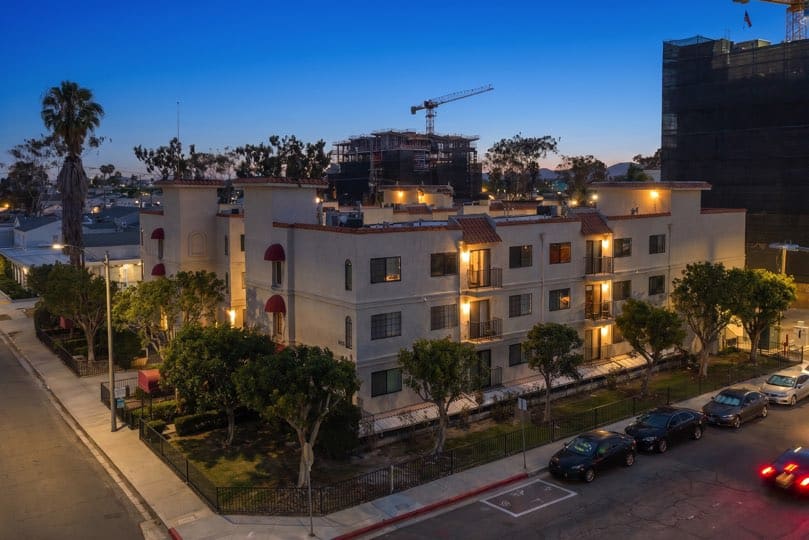 Exterior view of a multi-story stucco apartment building at dusk with lit windows, red awnings, surrounding trees, and cars parked along the street at 205 Ohio St Unit 304 in North Park.
