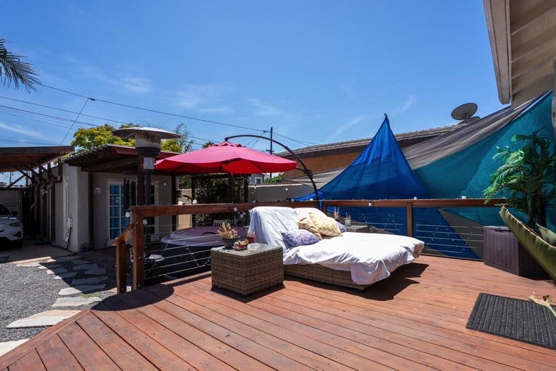 Wooden deck outdoor living space featuring a wicker lounge chair, red patio umbrella, hot tub, and blue shade sails at 3381 Bancroft St in North Park.