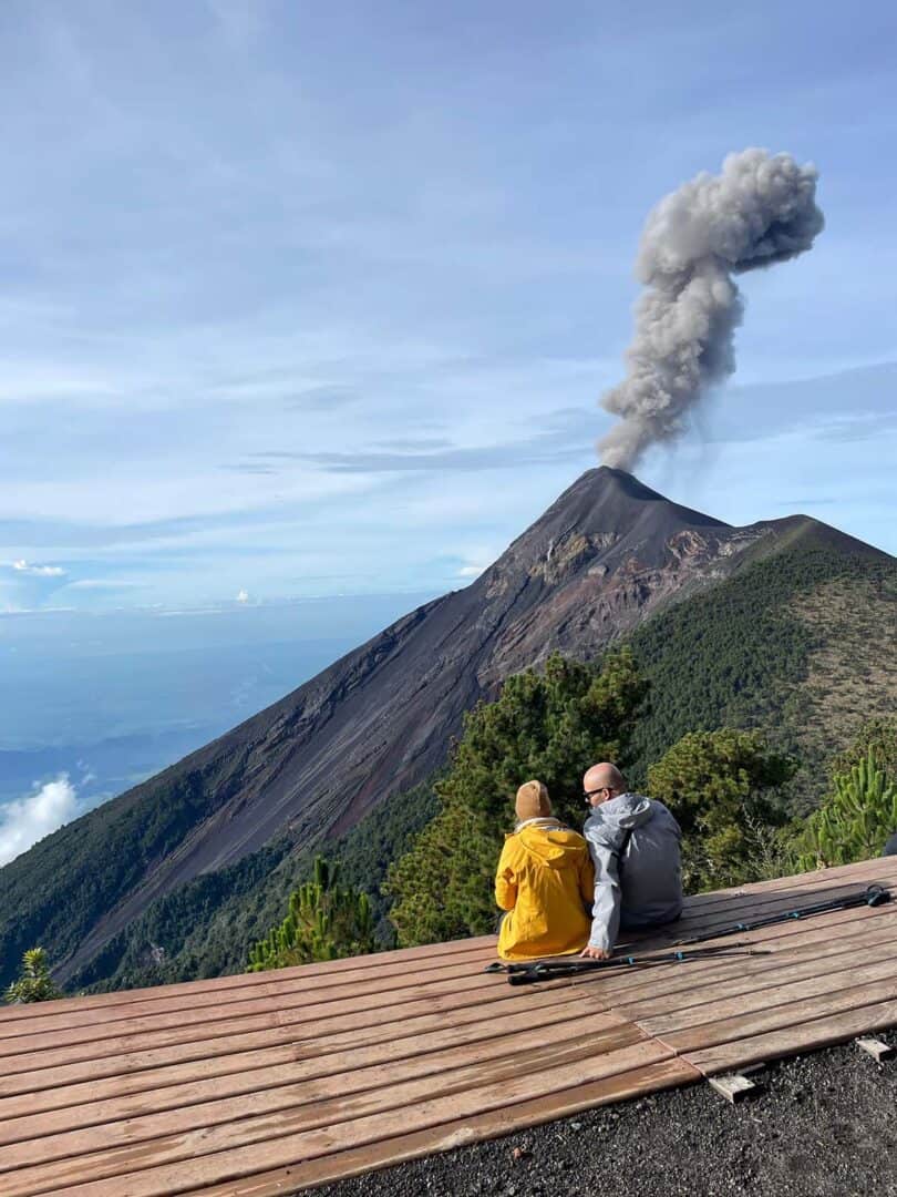 Two hikers seated on campsite platform watching an eruption of Fuego volcano from Acatenango